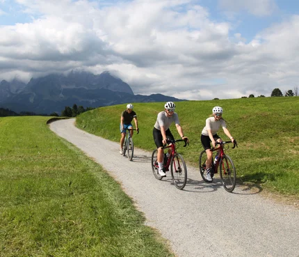 Drei Radfahrer auf Landstraße mit Bergen und Wolken im Hintergrund