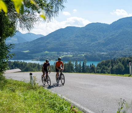 Zwei Radfahrer auf einer Landstraße mit Berg- und Seehintergrund