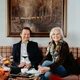 Zwei lächelnde Personen sitzen auf einem Sofa vor einem Waldgemälde