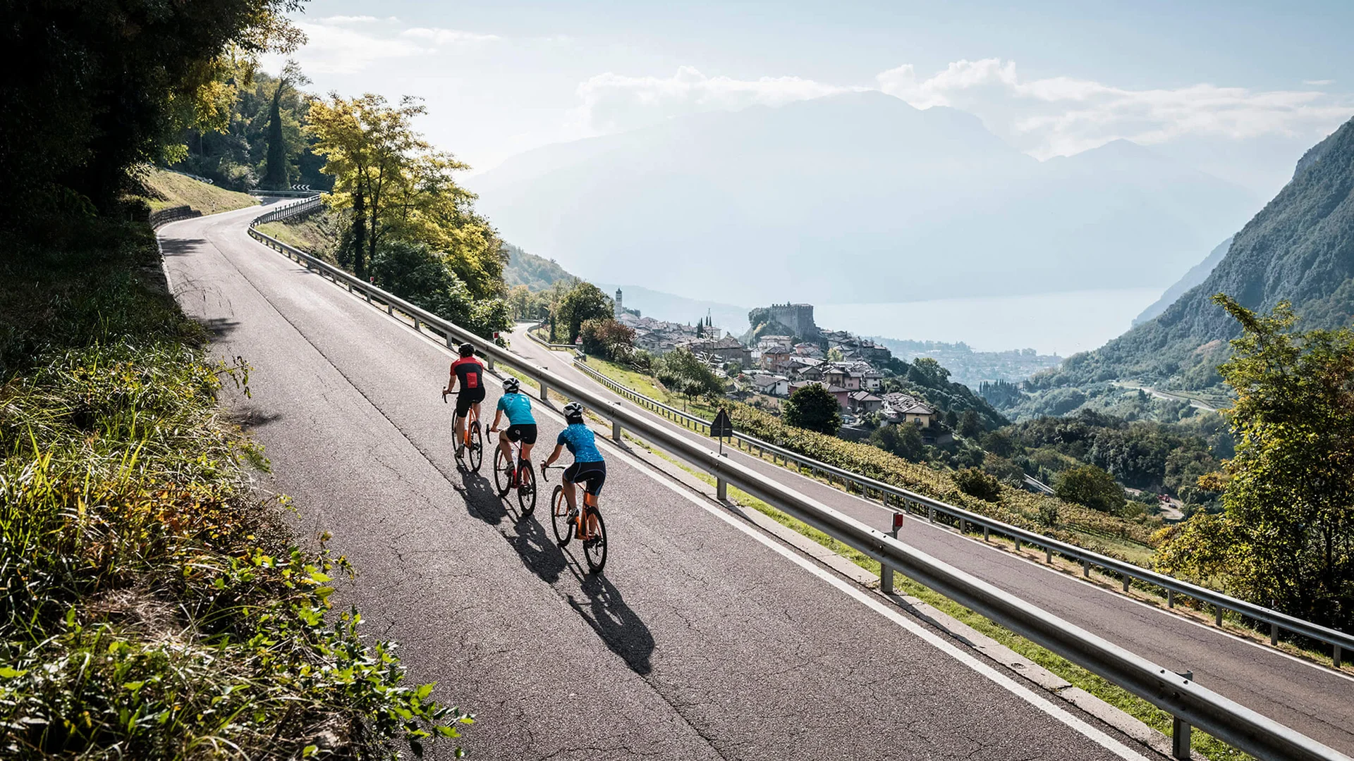 Roadbike Holidays © Alex Moling Drei Radfahrer fahren auf Bergstraße mit Blick auf Dorf und See