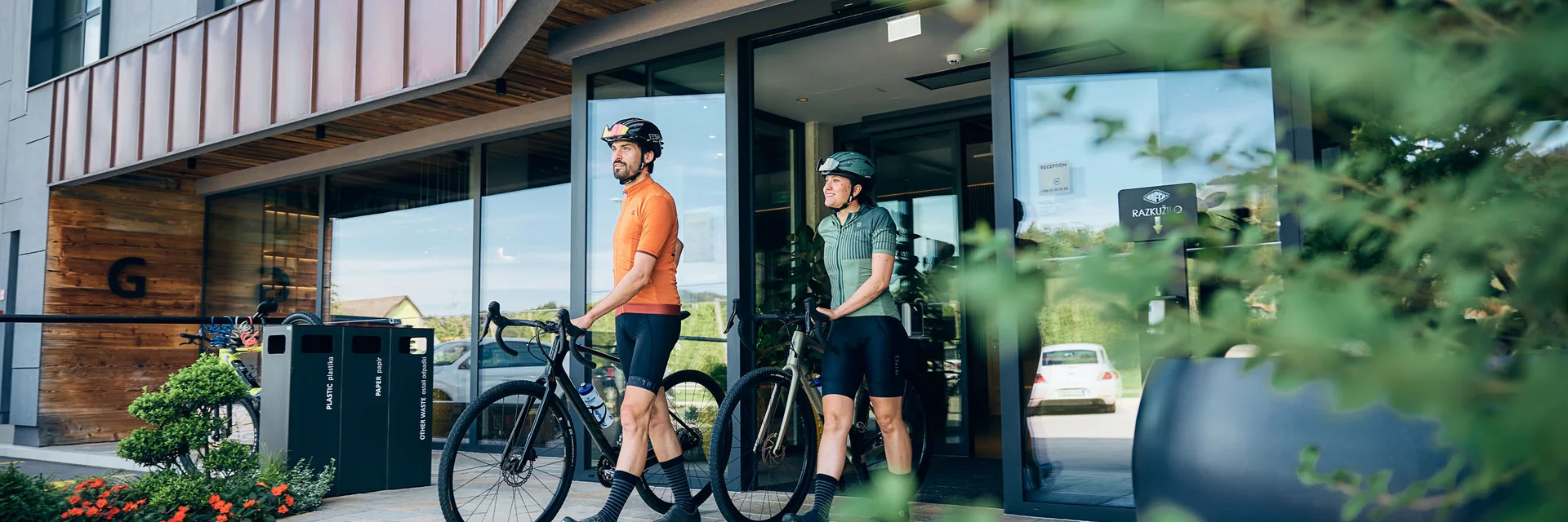 Gravelbike Holidays © Tobias Köhler Zwei Radfahrer in Sportkleidung gehen mit ihren Fahrrädern aus einem modernen Gebäude