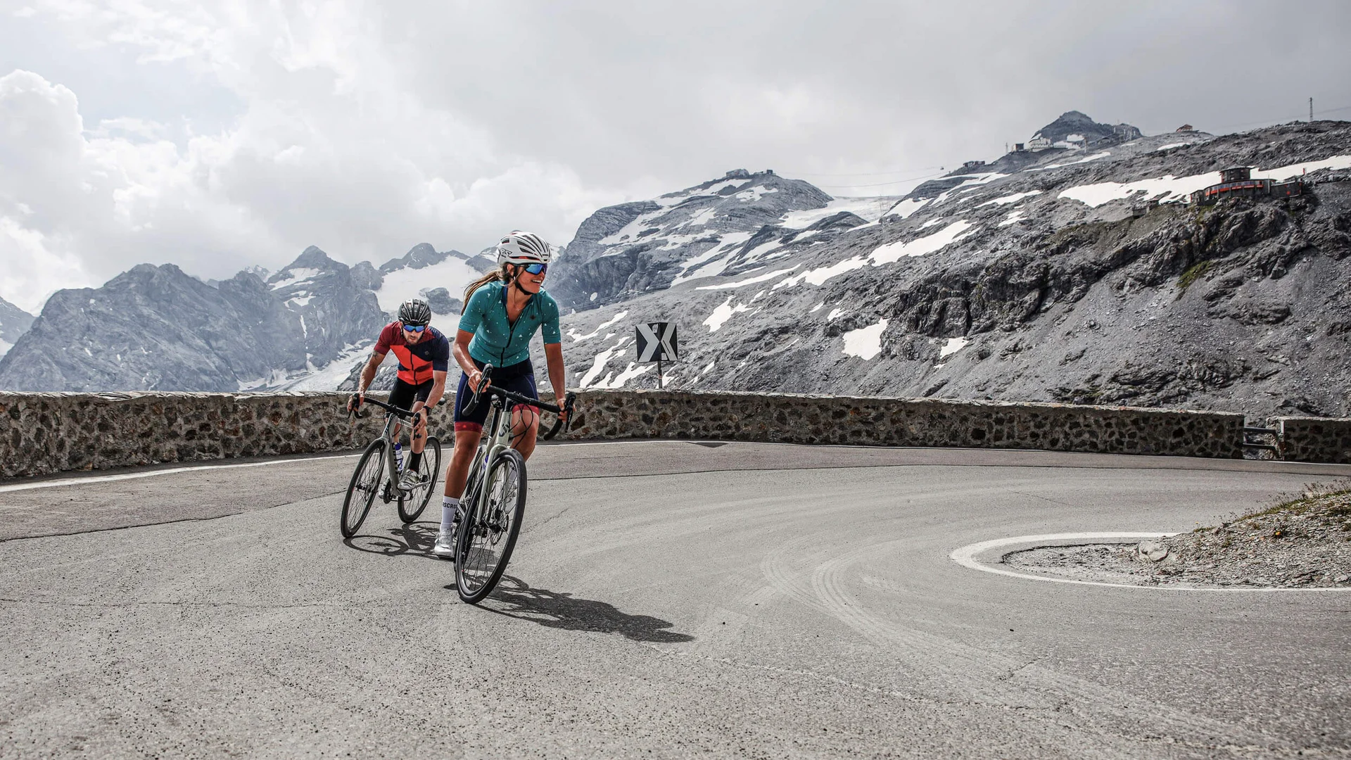 Bikehotel © Felix Saller Zwei Radfahrer fahren eine Bergstraße mit schneebedeckten Gipfeln im Hintergrund