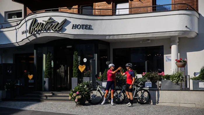 Zwei Radfahrer stoßen vor einem Hotel mit Blumenarrangements an.
