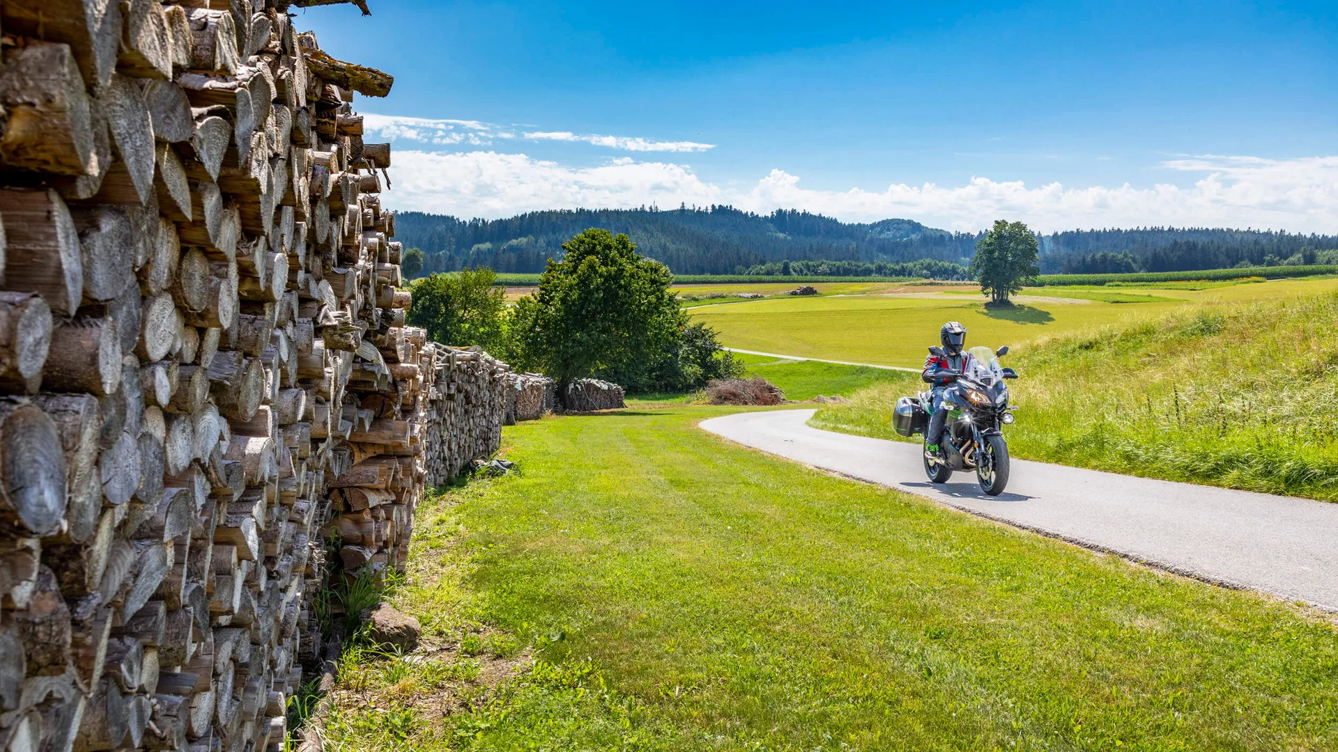 Motorradhotel © Alexander Seger Motorradfahrer auf Landstraße neben großem Holzstapel unter blauem Himmel