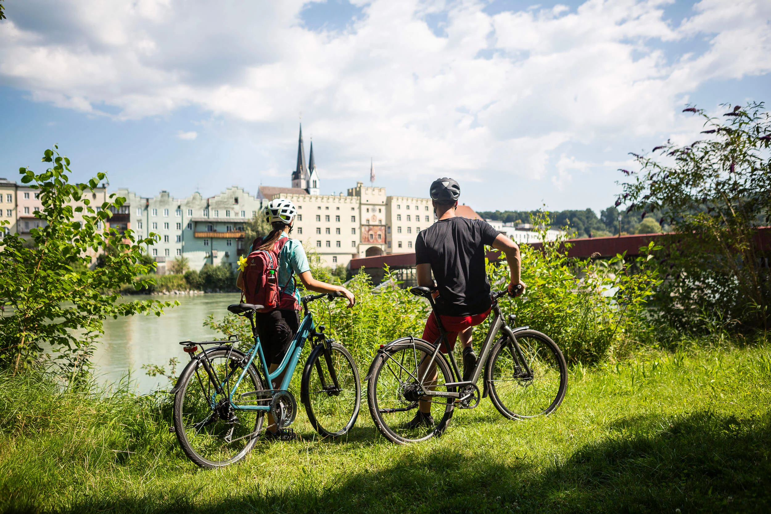 Zwei Radfahrer mit Helm blicken auf Gebäude am Flussufer an einem sonnigen Tag