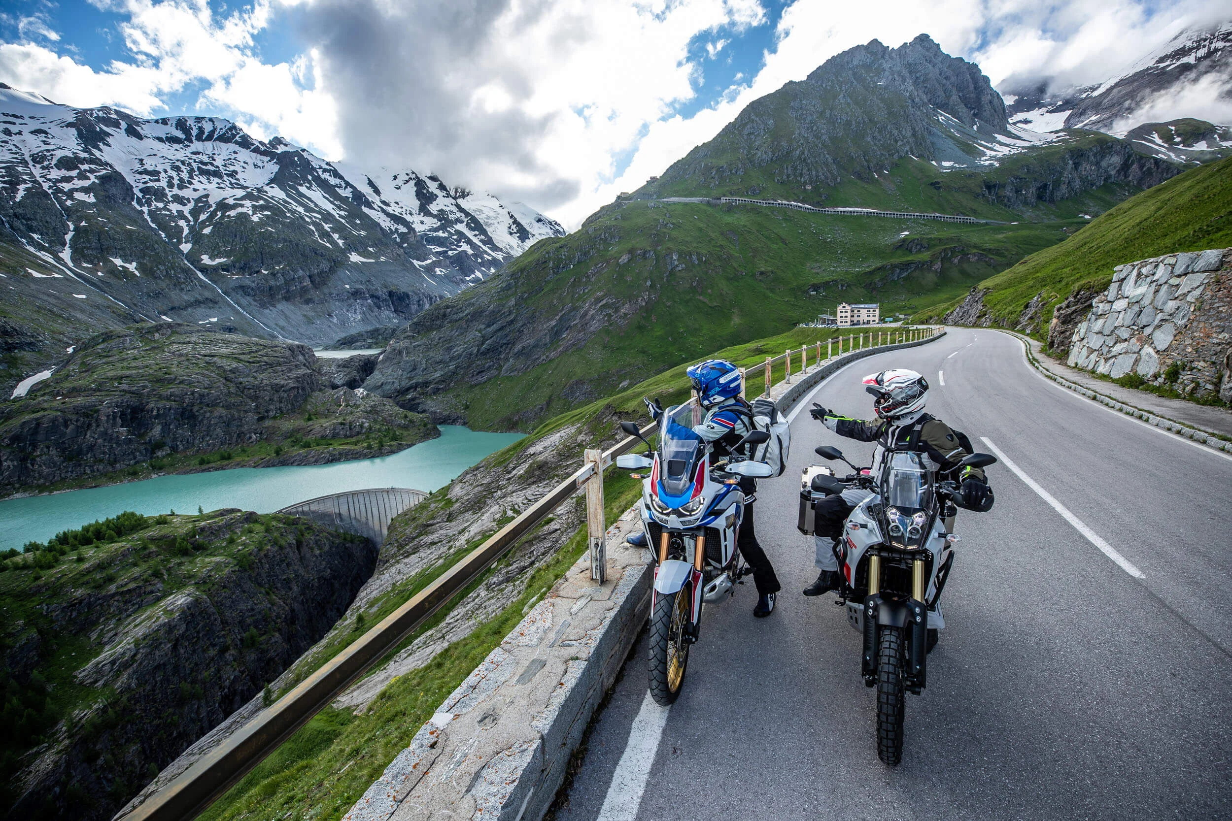 Motorradhotel © Arturo Rivas Zwei Motorradfahrer auf Alpenstraße mit Bergsee und schneebedeckten Gipfeln