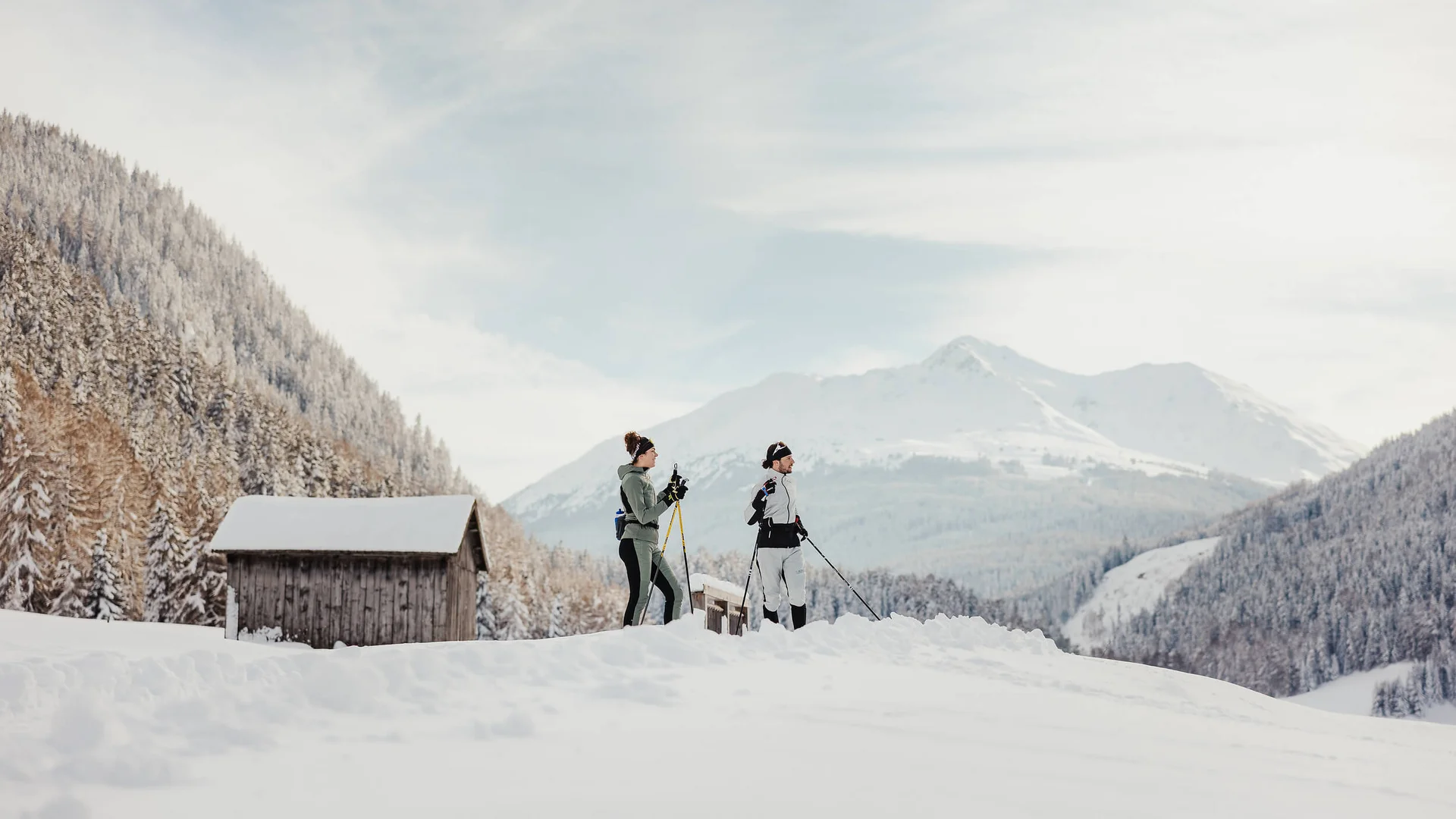 Zwei Personen beim Langlaufen im verschneiten Bergtal mit Hütten im Hintergrund