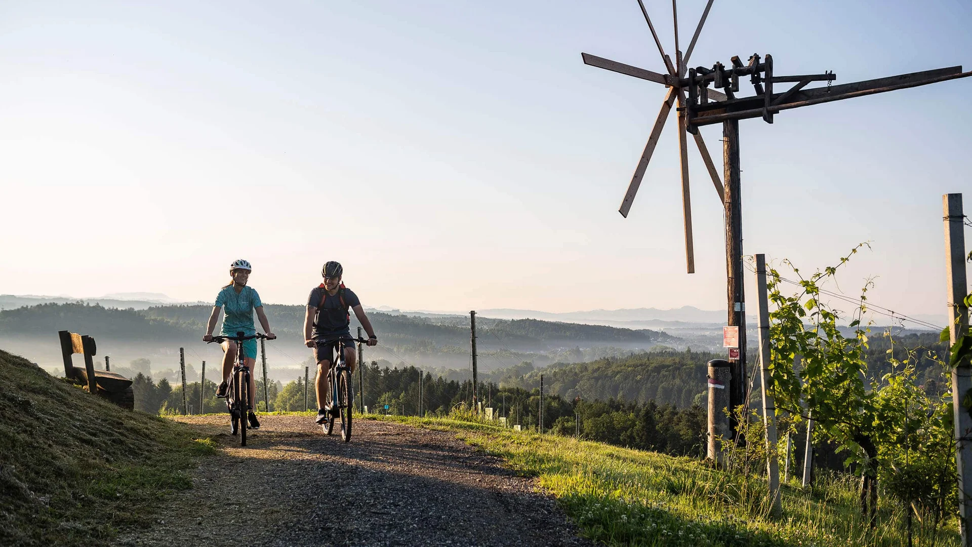 Zwei Radfahrer auf einem Landweg mit Windmühle und Hügelpanorama im Hintergrund
