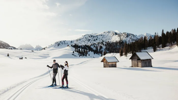 Langlauf & Skitourenhotel © Alex Moling Zwei Langläufer auf verschneiter Loipe mit Hütten und Bergen im Hintergrund