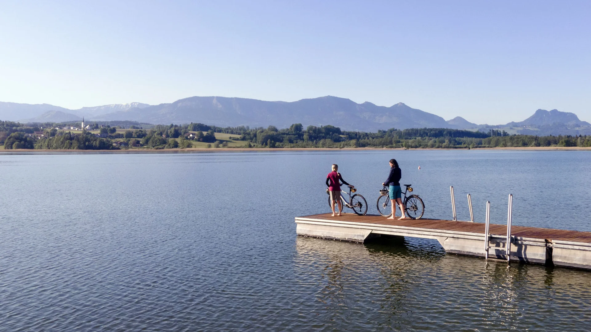 Radhotel © Oberbayern.de Zwei Personen mit Fahrrädern auf einem Steg an einem See mit Bergen im Hintergrund