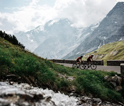 Zwei Radfahrer fahren auf Bergstraße mit schneebedeckten Bergen im Hintergrund