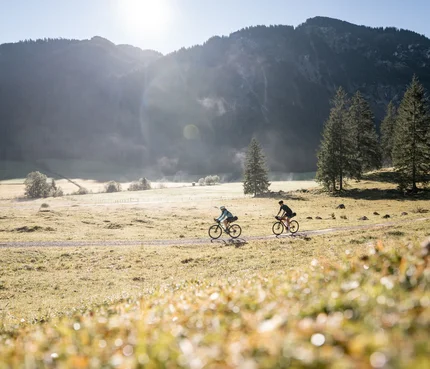 Zwei Radfahrer fahren auf einem Feldweg vor Bergkulisse bei Sonnenschein