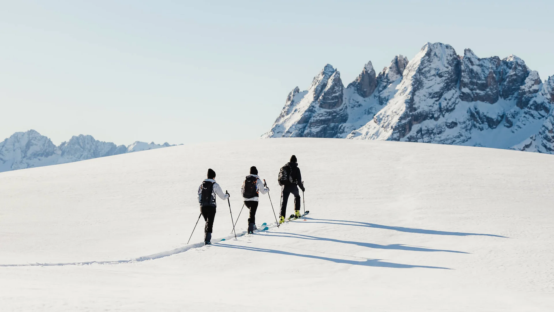 Drei Skifahrer wandern bei klarem Himmel auf schneebedecktem Hügel vor Bergkette