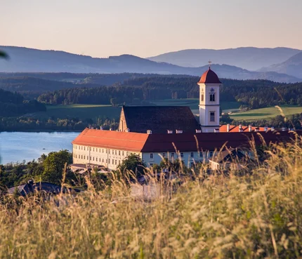 Klostergebäude mit rotem Dach vor Berg- und Seenlandschaft bei Sonnenuntergang