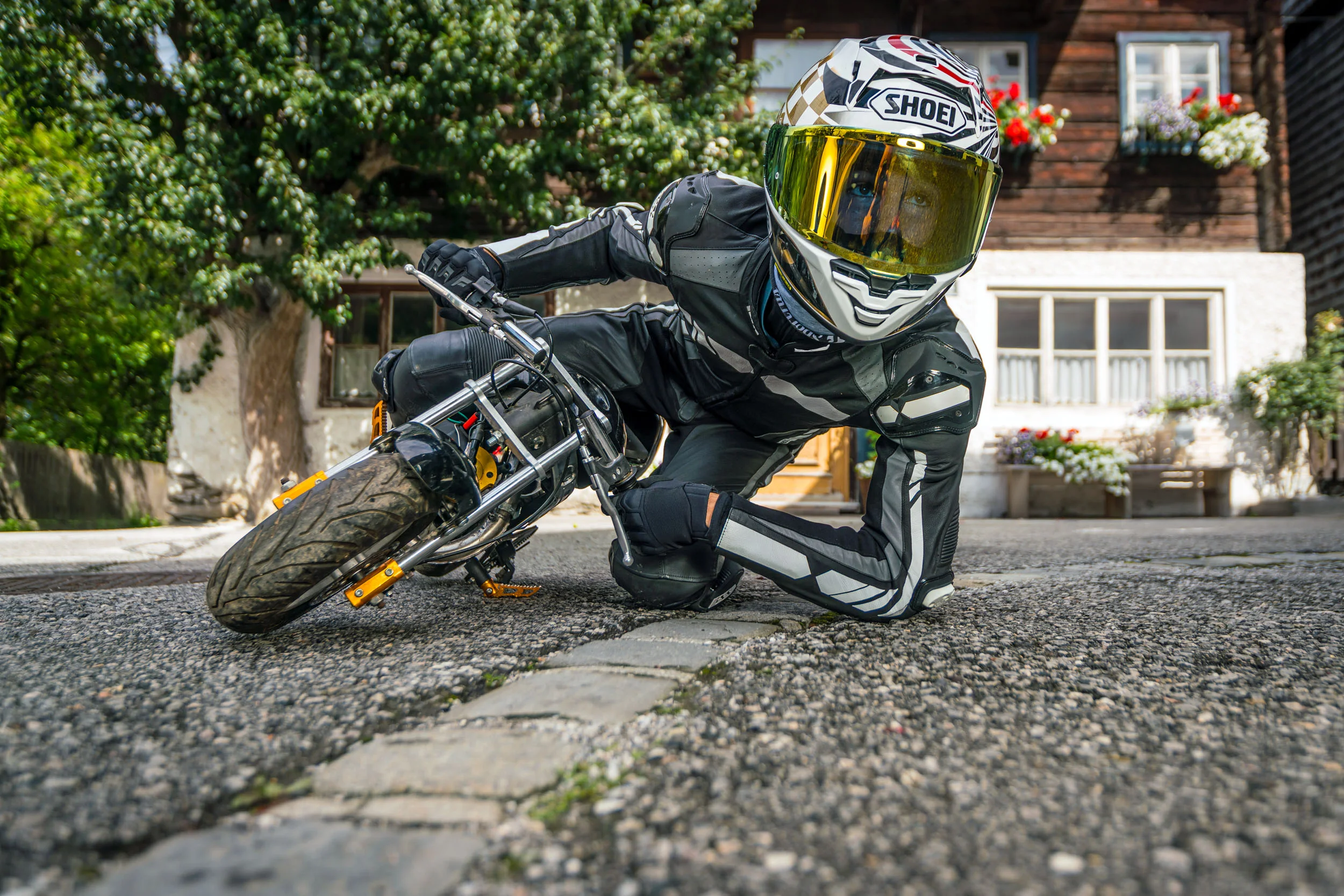 MoHo - Motorrad Hotels © Moppetfoto.de Person in Motorradkleidung liegt neben kleinem Motorrad auf Straße vor Haus