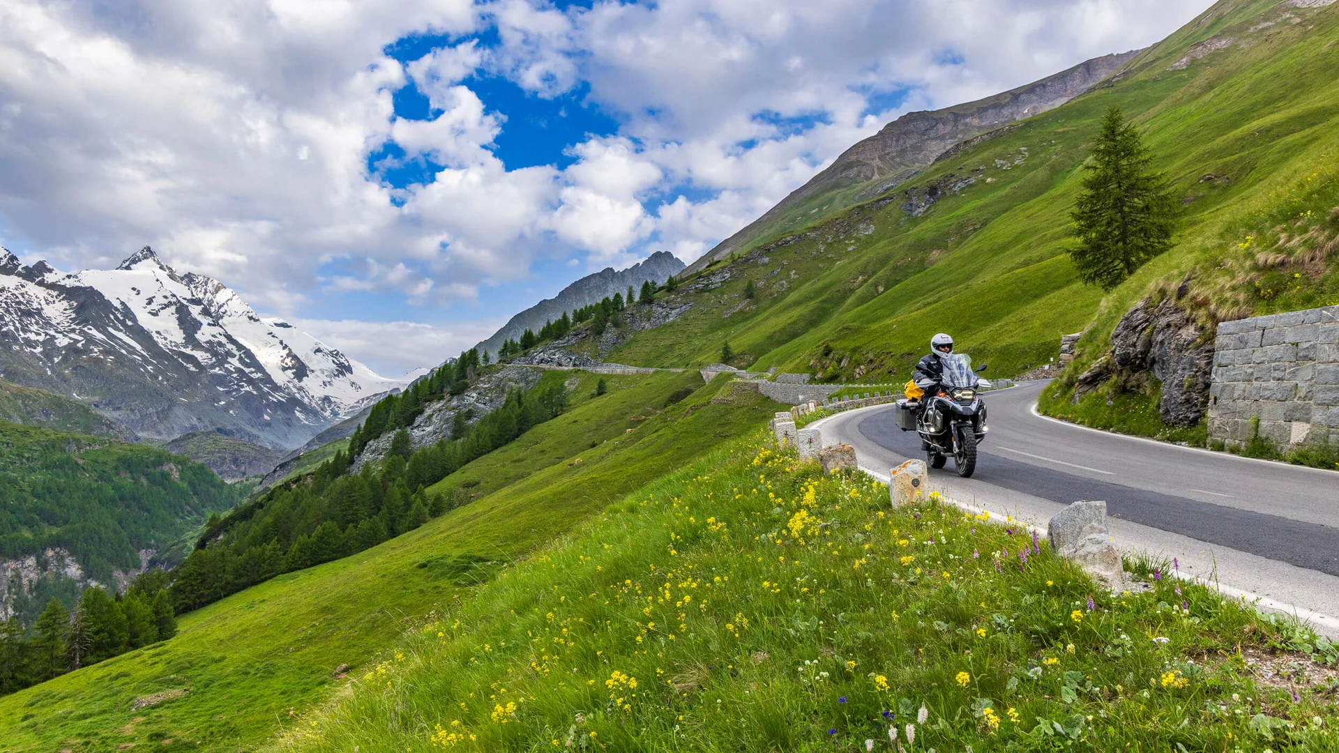 MoHo - Motorrad Hotels © Alexander Seger Motorradfahrer auf kurvenreicher Bergstraße mit grünen Wiesen und schneebedeckten Gipfeln