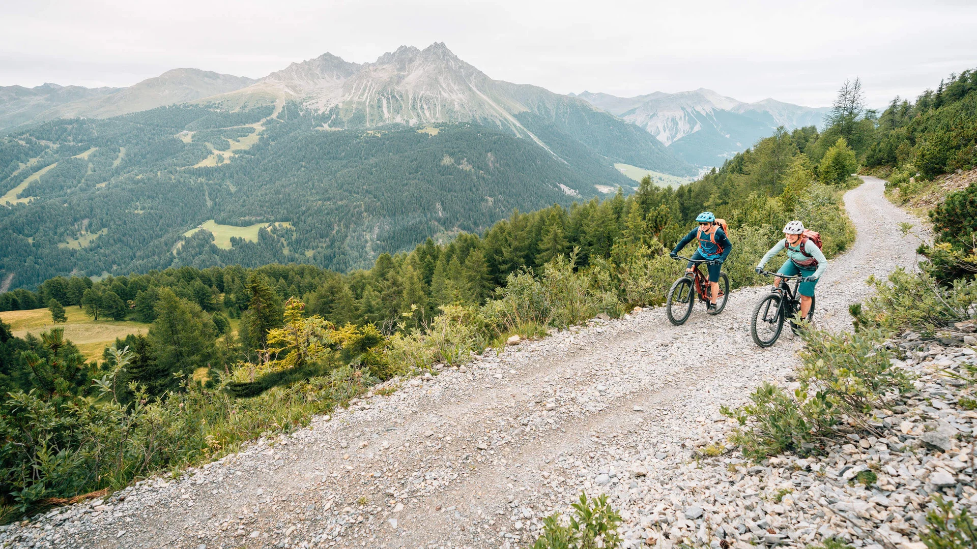 Zwei Radfahrer fahren auf einem Bergweg mit Alpen im Hintergrund