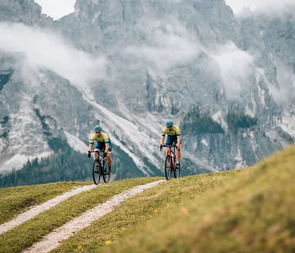 Zwei Radfahrer fahren auf einem Bergweg vor nebligen Alpenfelsen